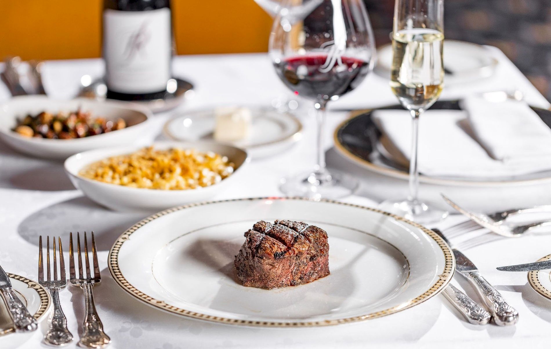 A fine dining table with a small steak on a white plate, surrounded by wine glasses, bowls of sides, and a bottle in the background.