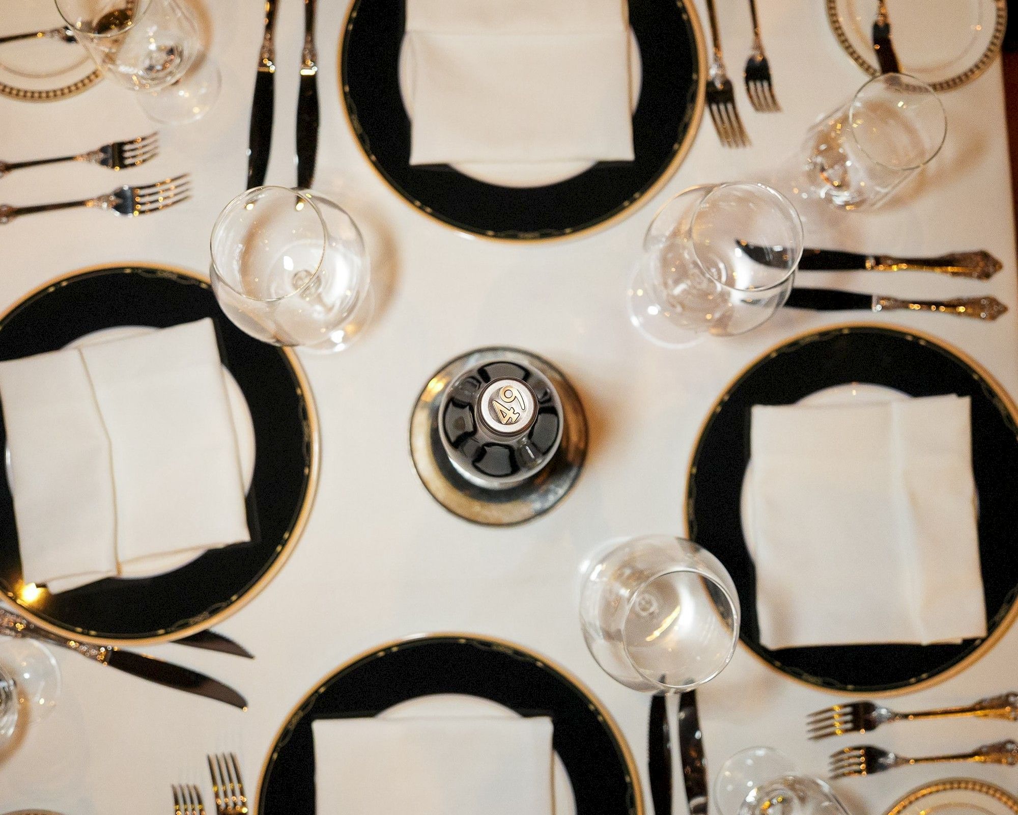 A formal dining table set with black-edged plates, white napkins, multiple forks and knives, and a central small round centerpiece.
