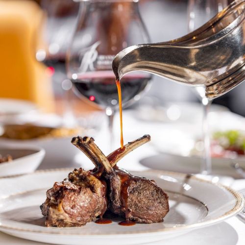 Tender lamb chops with bones on a plate, a glossy sauce being poured over them, and wine glasses in a fine dining setting. End with period.