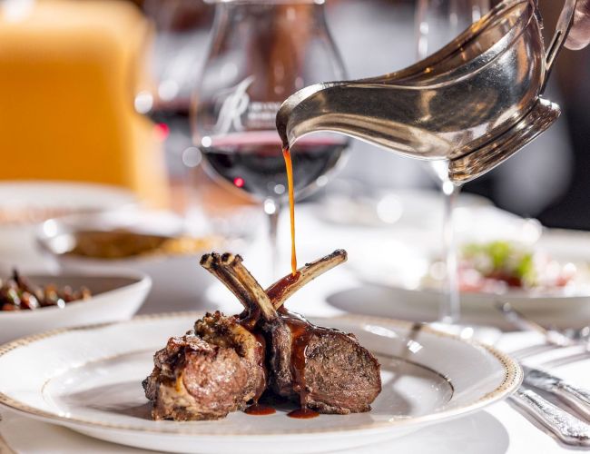 Tender lamb chops with bones on a plate, a glossy sauce being poured over them, and wine glasses in a fine dining setting. End with period.