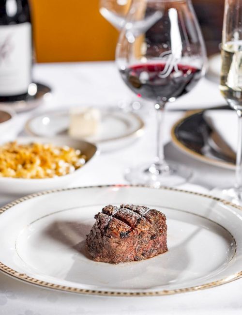 A fine dining table with a plated steak centerpiece, wine glasses, bread bowls, and side dishes in a classy setting. Ending with a period.