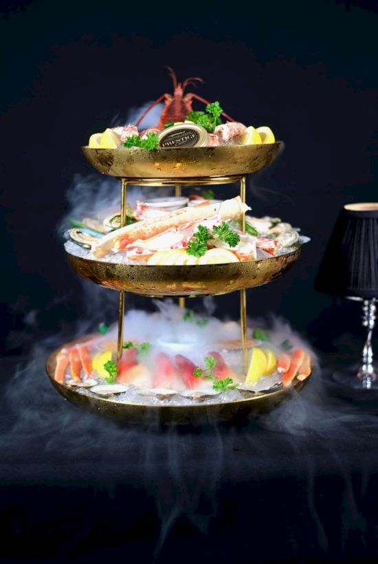 A three-tiered hot pot or seafood platter with prawns and greens, steaming over a smoky, dark background.