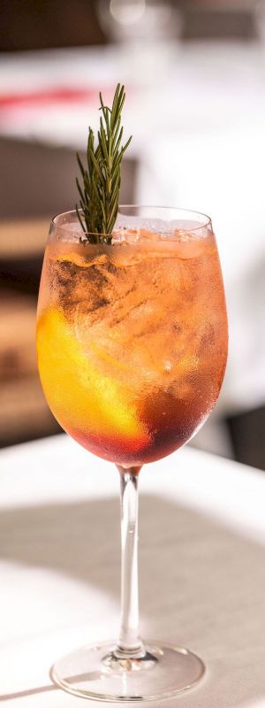 A cocktail in a wine glass with ice and orange slices, garnished with a sprig of rosemary, on a sunny table.