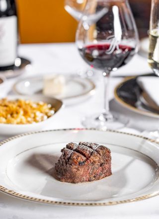 A fine dining table with a small steak or beef fillet on a plate, surrounded by wine glasses, bread, side dishes, and elegant cutlery, ready to be served.