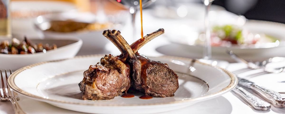 A plate with lamb chops, sauce being poured over them, and a refined dining setup in the background.